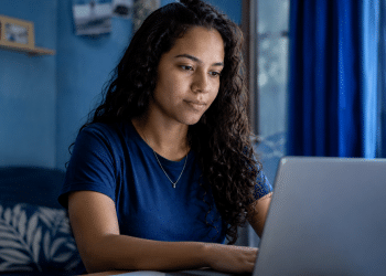 Jovem de cabelos cacheados usando camiseta azul trabalha em notebook próximo à janela com cortinas azuis.