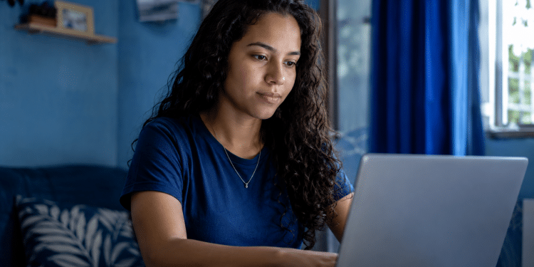 Jovem de cabelos cacheados usando camiseta azul trabalha em notebook próximo à janela com cortinas azuis.