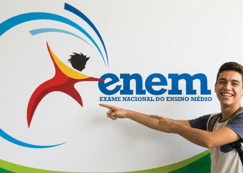 Jovem sorridente aponta para o logotipo do Enem fixado em parede branca