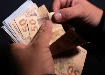 Pessoa conta notas de cinquenta reais segurando cédulas brasileiras em fundo escuro, simbolizando o pagamento de R$ 2.821 nos dias 1, 2 e 3 de julho.