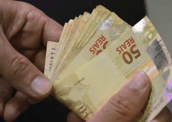 Mãos segurando maço de cédulas de R$ 50 reais brasileiros em fundo escuro, representando pagamentos, simbolizando os depósitos de junho do INSS.