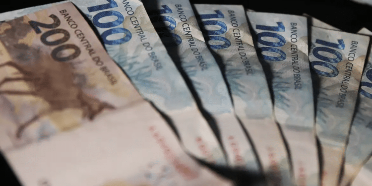 Cédulas de real de cem e duzentos reais dispostas em leque sobre fundo escuro, simbolizando o pagamento de junho sem o adicional de R$ 810,50.