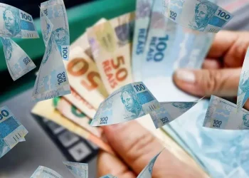 Mãos segurando dinheiro em espécie sobre notas de reais, representando o pagamento de R$ 1.450 do Pé-de-Meia que está prestes a ser iniciado.
