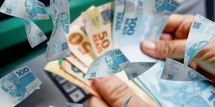 Mãos segurando dinheiro em espécie sobre notas de reais, representando o pagamento de R$ 1.450 do Pé-de-Meia que está prestes a ser iniciado.