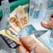 Mãos segurando dinheiro em espécie sobre notas de reais, representando o pagamento de R$ 1.450 do Pé-de-Meia que está prestes a ser iniciado.
