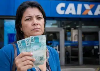 Mulher segura notas de 100 reais em frente a uma agência da Caixa, representando o pagamento tardio de R$ 1.621 para CPFs com finais de 0 a 9.