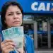 Mulher segura notas de 100 reais em frente a uma agência da Caixa, representando o pagamento tardio de R$ 1.621 para CPFs com finais de 0 a 9.