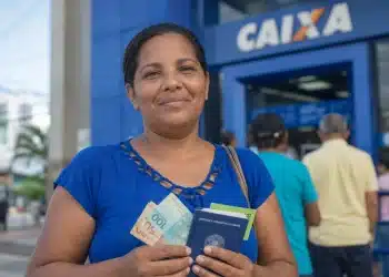 Mulher segura cédulas de reais e documentos em frente a uma agência da Caixa, representando o pagamento unificado do Bolsa Família em junho.