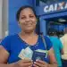 Mulher segura cédulas de reais e documentos em frente a uma agência da Caixa, representando o pagamento unificado do Bolsa Família em junho.