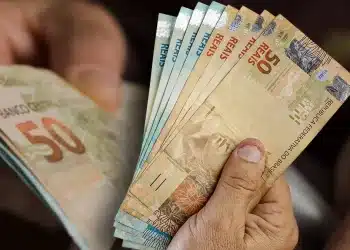 Mão segurando cédulas de real em destaque, representando pagamentos de R$ 946, R$ 1.216 e R$ 1.486 confirmados pela CAIXA.