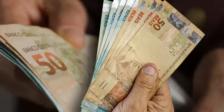 Mão segurando cédulas de real em destaque, representando pagamentos de R$ 946, R$ 1.216 e R$ 1.486 confirmados pela CAIXA.