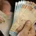 Mão segurando cédulas de real em destaque, representando pagamentos de R$ 946, R$ 1.216 e R$ 1.486 confirmados pela CAIXA.