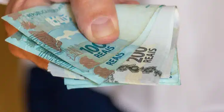 Mão segurando cédulas de real, representando pagamentos do INSS, abono PIS/Pasep e ressarcimentos liberados antes de julho.