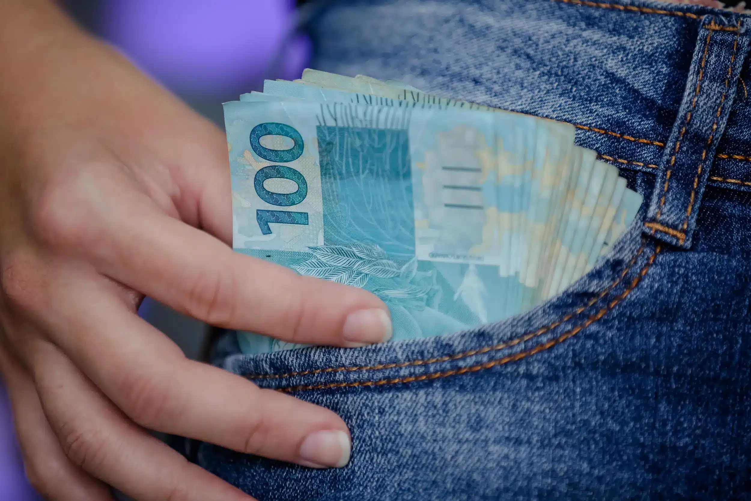 Notas de R$ 100 guardadas no bolso de uma calça jeans, simbolizando atenção ao calendário de pagamentos de junho.