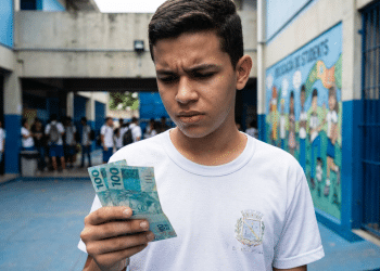 Estudante de uniforme branco observa cédulas de R$ 100 no pátio da escola, representando o incentivo do Pé-de-Meia para quem faz o Enem.
