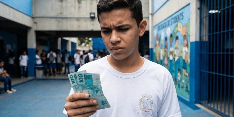 Estudante de uniforme branco observa cédulas de R$ 100 no pátio da escola, representando o incentivo do Pé-de-Meia para quem faz o Enem.