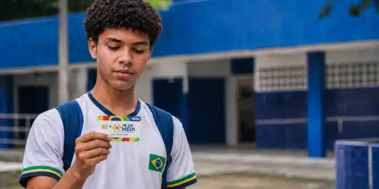 Estudante segura cartão do Pé-de-Meia em frente a uma escola pública, representando a dúvida sobre quem recebe o benefício em junho.