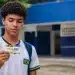 Estudante segura cartão do Pé-de-Meia em frente a uma escola pública, representando a dúvida sobre quem recebe o benefício em junho.