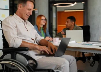 Profissional com deficiência em cadeira de rodas utilizando notebook em ambiente corporativo moderno, com colegas ao fundo em reunião colaborativa.