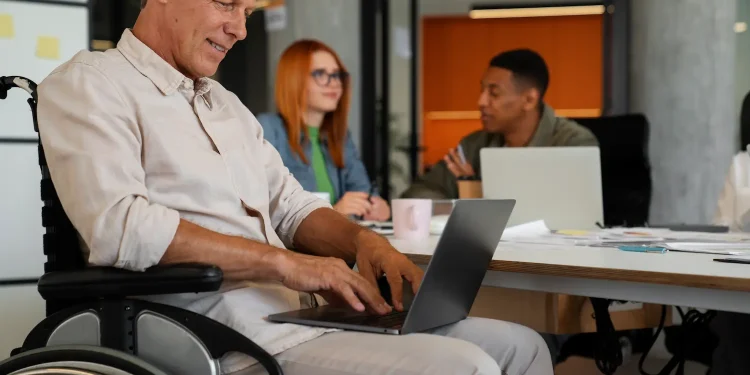 Profissional com deficiência em cadeira de rodas utilizando notebook em ambiente corporativo moderno, com colegas ao fundo em reunião colaborativa.
