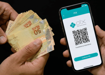 Mãos segurando notas de real e celular exibindo QR Code do PIX Banco Central, simbolizando o Pix automático de R$ 1.000 do Imposto de Renda.