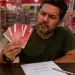Homem com expressão de dúvida observa vales-compra vermelhos em uma loja, ao lado de uma folha de anotações com diferentes formas de escrita da palavra, representando a dúvida sobre o plural de vale-compra.