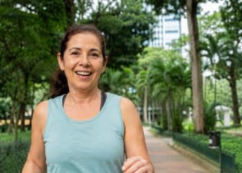 Mulher madura sorri ao caminhar em um parque arborizado, unindo exercício e contato com a natureza, dois hábitos do envelhecer bem.