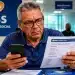 Idoso em agência do INSS analisando documento de extrato de pagamento enquanto verifica no celular informações sobre descontos indevidos e alerta de golpes