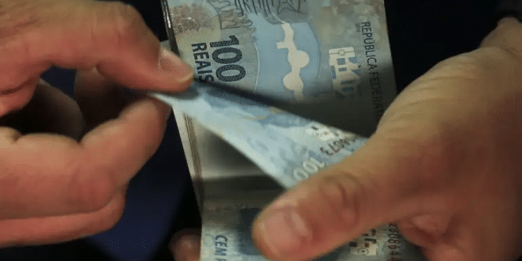 Mão segura cédulas de cem reais em leque diante da fachada de uma agência da Caixa