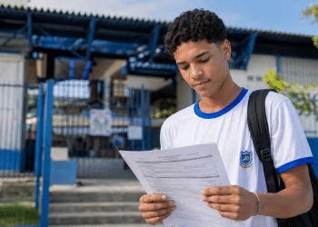Estudante jovem lendo documento de inscrição em frente a uma escola pública, representando processo seletivo do Instituto Federal.