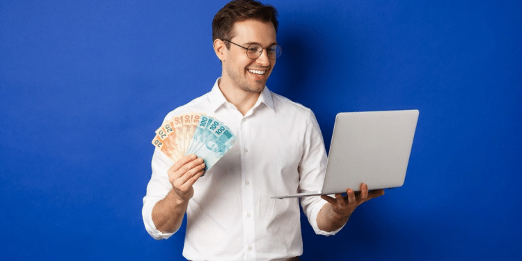 Homem sorrindo segura notas de real ao lado de notebook aberto em fundo azul.