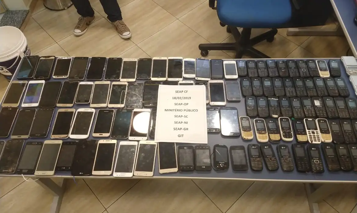 Mesa coberta por dezenas de celulares apreendidos de vários modelos, com uma ficha de identificação policial ao centro.