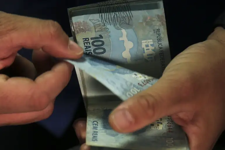 Pessoa conta cédulas de cem reais com as duas mãos, simbolizando o pagamento do Abono Salarial 2026.