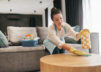 Mulher de luvas limpa a superfície de uma mesa na sala, com balde de produtos de limpeza ao lado