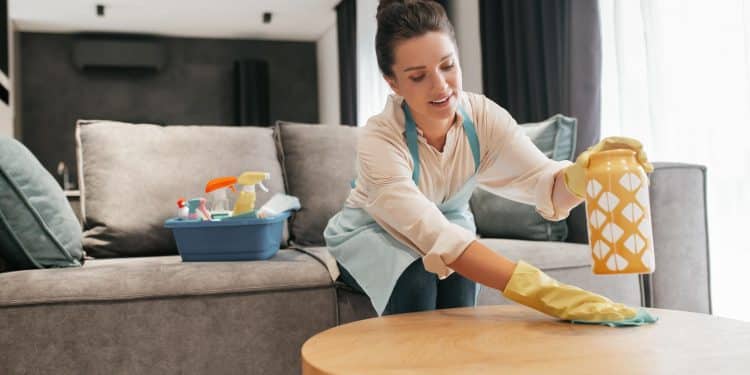 Mulher de luvas limpa a superfície de uma mesa na sala, com balde de produtos de limpeza ao lado