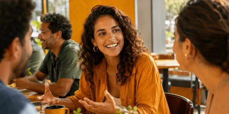 Jovem brasileira sorrindo e conversando com outras pessoas em cafeteria, representando empatia e habilidade social.