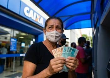 Mulher segura notas de R$ 50 e R$ 100 em frente a uma agência da Caixa, em imagem que ilustra os dois pagamentos com prazo em 20 de junho: o Abono Salarial e o ressarcimento do INSS