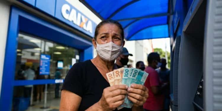 Mulher segura notas de R$ 50 e R$ 100 em frente a uma agência da Caixa, em imagem que ilustra os dois pagamentos com prazo em 20 de junho: o Abono Salarial e o ressarcimento do INSS
