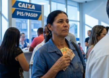 Mulher segura notas de dinheiro em uma fila dentro de uma agência da Caixa, sob um letreiro escrito PIS/Pasep, representando o pagamento do ressarcimento.
