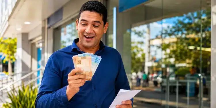 Servidor público sorrindo ao segurar dinheiro e envelope de pagamento em frente a uma agência bancária.