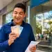 Servidor público sorrindo ao segurar dinheiro e envelope de pagamento em frente a uma agência bancária.