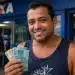 Homem sorridente segura cédulas de real em frente a uma agência da Caixa, representando o Saque Calamidade de até R$ 6.220.