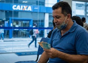 Homem conferindo cédulas de R$ 100 e R$ 50 em frente a uma agência da Caixa, referência ao Saque Calamidade do FGTS