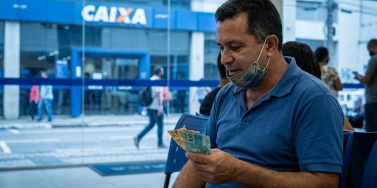 Homem conferindo cédulas de R$ 100 e R$ 50 em frente a uma agência da Caixa, referência ao Saque Calamidade do FGTS