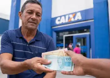 Trabalhador recebendo cédulas de real em frente a uma agência da Caixa, representando o saque de até R$ 1.621 do PIS/Pasep.