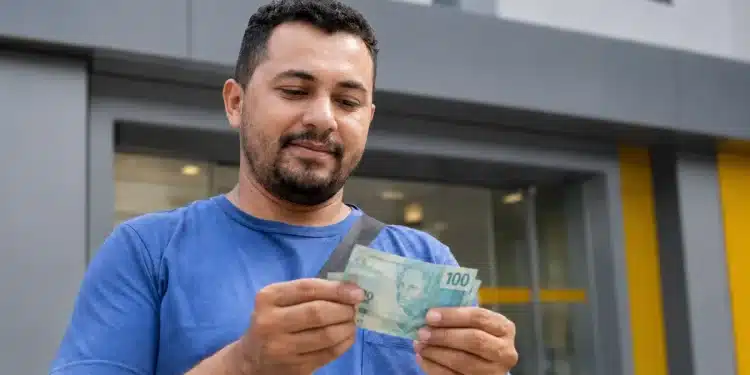 Homem segurando dinheiro em frente a uma agência bancária, ilustrando pagamento da restituição do segundo lote do Imposto de Renda