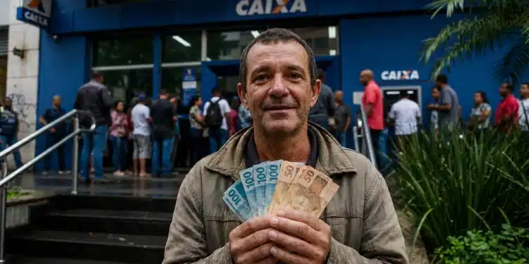 Homem segura notas de dinheiro em frente a uma agência da Caixa, em referência ao pagamento do seguro-desemprego em 2026.