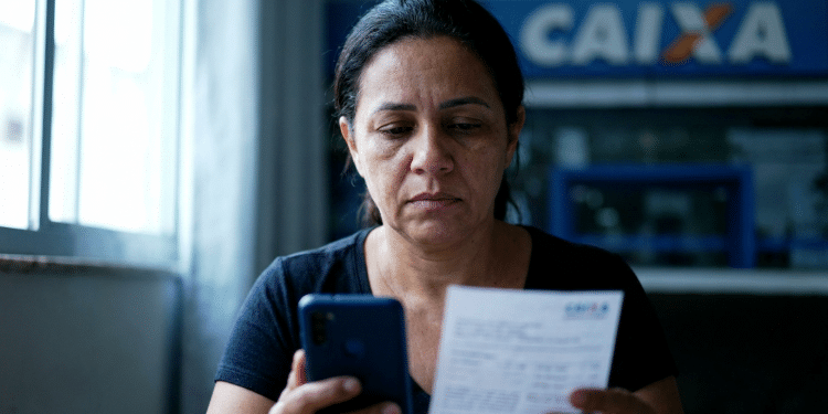 Mulher confere documento e celular em frente a uma agência da Caixa sobre o ressarcimento do PIS/Pasep.