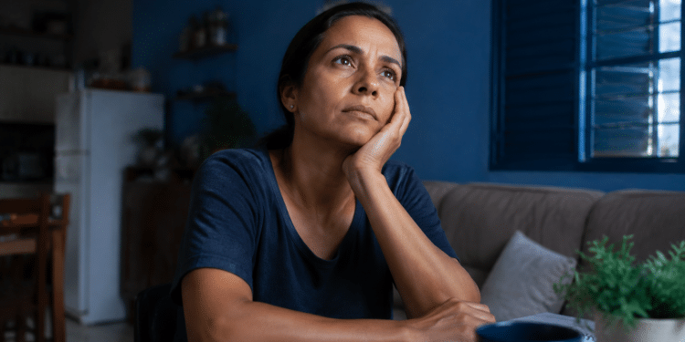 Mulher em ambiente doméstico com expressão de preocupação refletindo sobre sobrecarga mental e ansiedade.