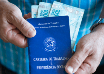 Mãos seguram Carteira de Trabalho azul e notas de 100 reais em roupa xadrez azul.